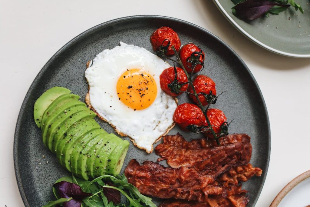 A delicious keto breakfast featuring fried egg, bacon, avocado, and tomatoes on a plate.